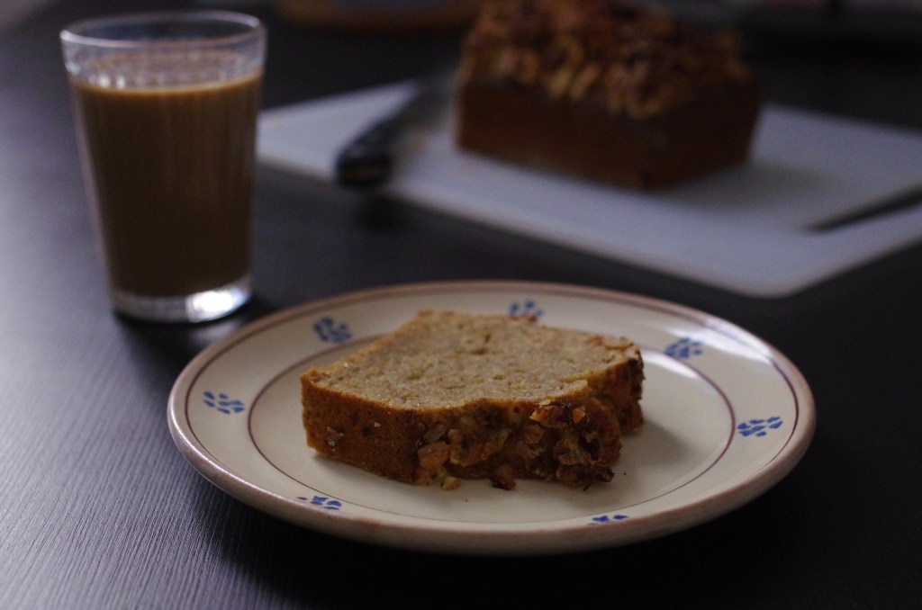 Fetta di Banana Bread in primo piano, accompagnato da caffè e latte. Sullo sfondo, suonato, il Banana Bread sul tagliere.